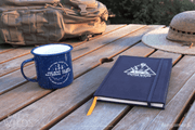 A photograph of two new Outer Wilds items on a weathered wooden table, with a camping pack and a hat at the edge of the frame. A navy Outer Wilds camping mug says 